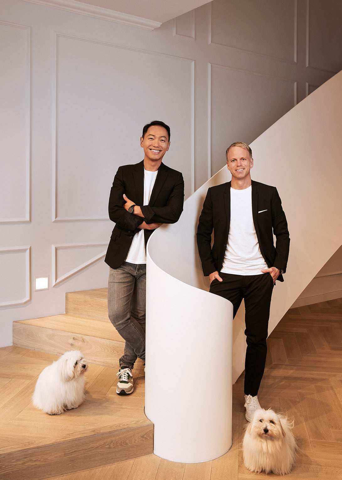 Budi and Philipp standing on a staircase in their atelier reception with two Coton de Tulear dogs
